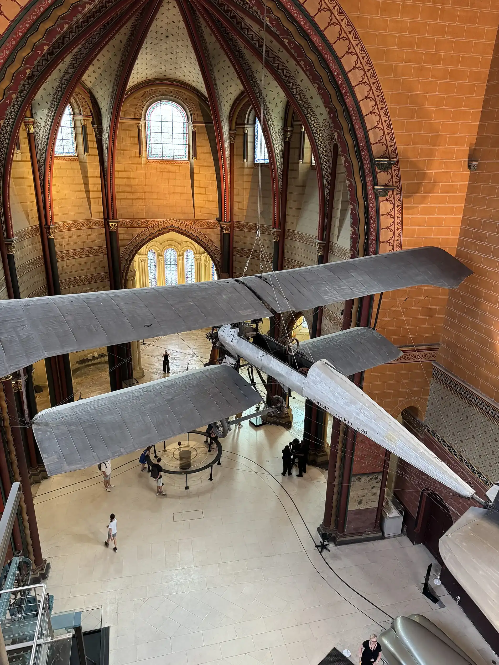 Early Breguet Type R.U1 aircraft suspended beneath the painted vaulted ceiling of the priory church at the Musée des Arts et Métiers, with visitors and the Foucault pendulum circle on the floor below