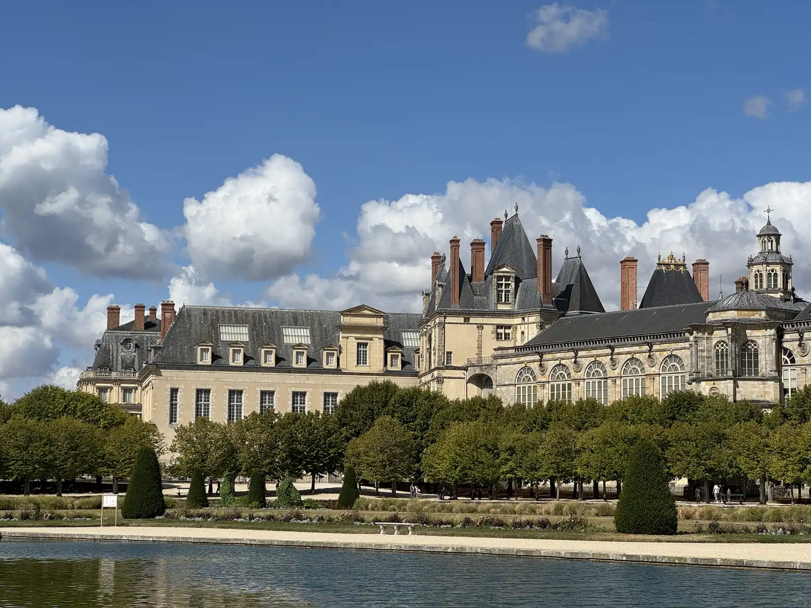 Rooftops and chimneys of the Château de Fontainebleau behind clipped trees, with a bench by the pond
